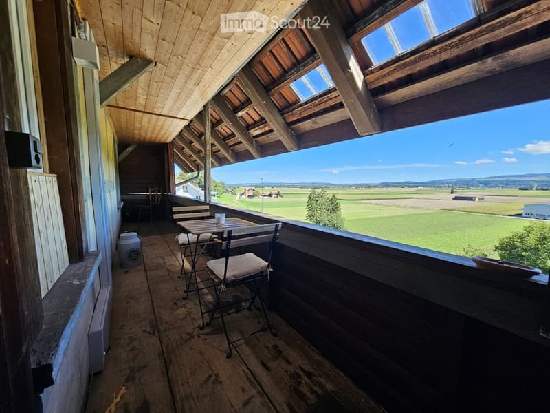 Wooden balcony with slatted ceiling, table, chairs, cup, and barrel, overlooking a green field.