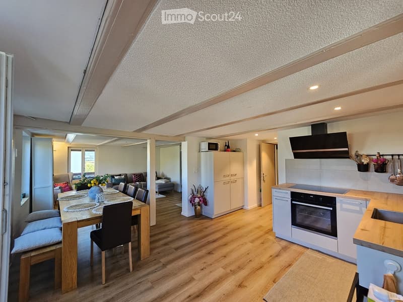 Open-plan living and dining area with wooden flooring, a dining table with chairs, a sofa, and a modern kitchen with white cabinets, an oven, and a microwave.