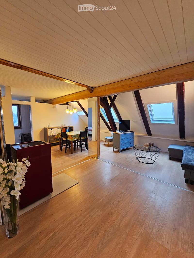 Open-plan living and dining area with wooden floors, exposed wooden beams, large windows, and modern furniture including a sofa, coffee table, dining table, and chairs.