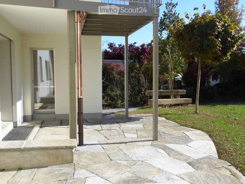 Outdoor patio area with stone paving, covered walkway, grassy lawn, trees, and a wooden bench.