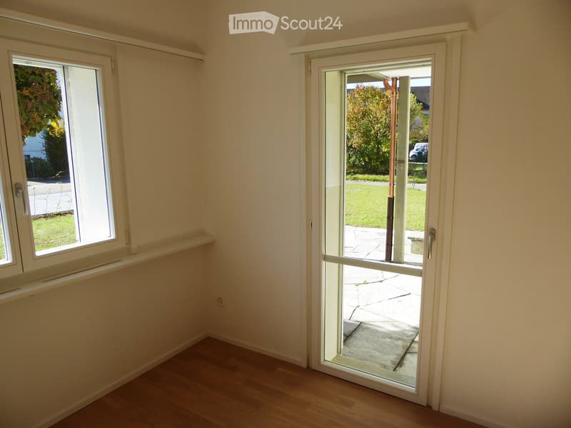 Empty room with wooden flooring, white walls, large window, and open sliding glass door leading to a patio area.