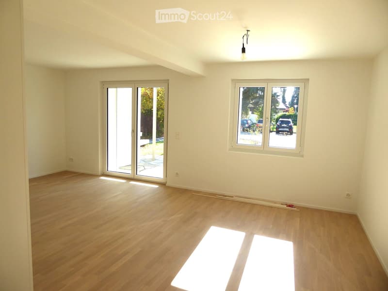 Empty room with light wooden flooring, white walls, large windows, and a sliding glass door leading outside.