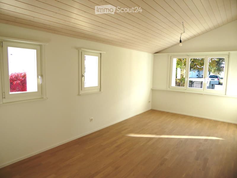 Empty room with light wooden flooring, white walls, and multiple windows offering natural light. The ceiling has a slanted design with visible wooden paneling.