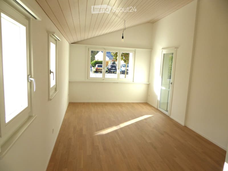 Empty room with light wooden flooring, white walls, and large windows allowing natural light. The room features a sliding glass door leading outside and a slanted ceiling with exposed wooden beams.