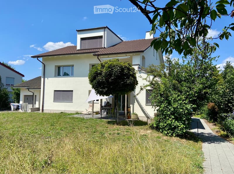 Two-story white house with a tiled roof, surrounded by a well-maintained garden with trees and outdoor furniture.