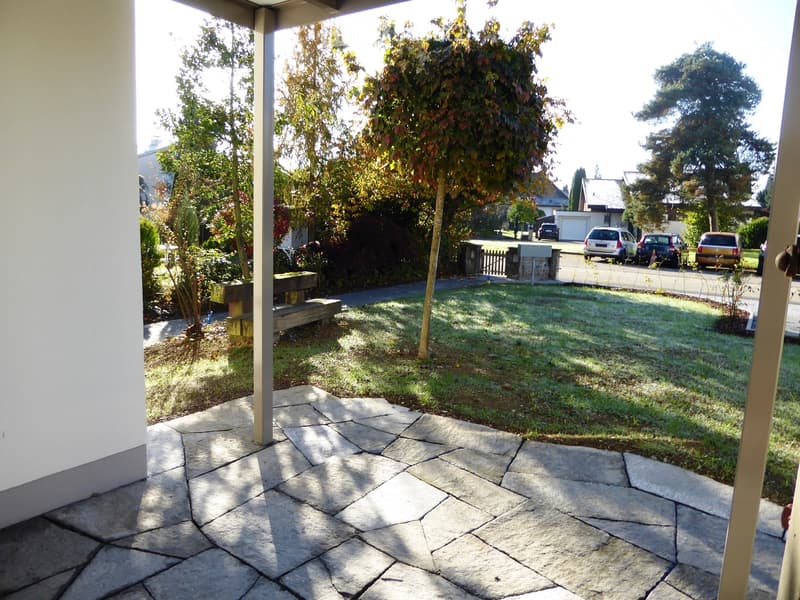 Garden area with a stone-paved pathway, grassy lawn, trees, and a bench near the house.