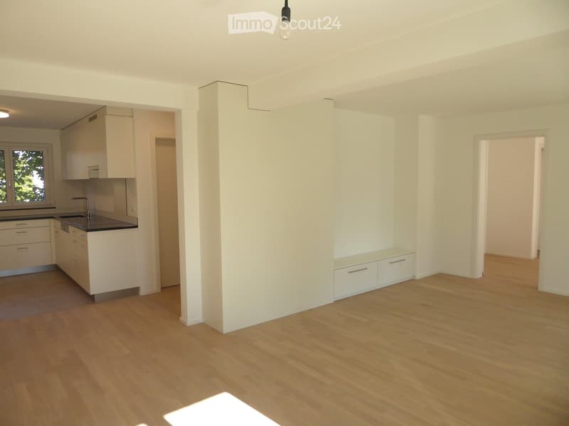 Empty room with light wooden flooring, white walls, and a small kitchen area visible in the background. No furniture or personal items are present.