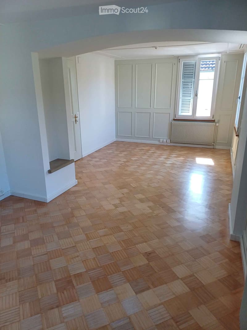Empty room with wooden herringbone flooring, white walls, and large windows with shutters. A radiator is visible under one window, and a small bench is placed near the left wall.