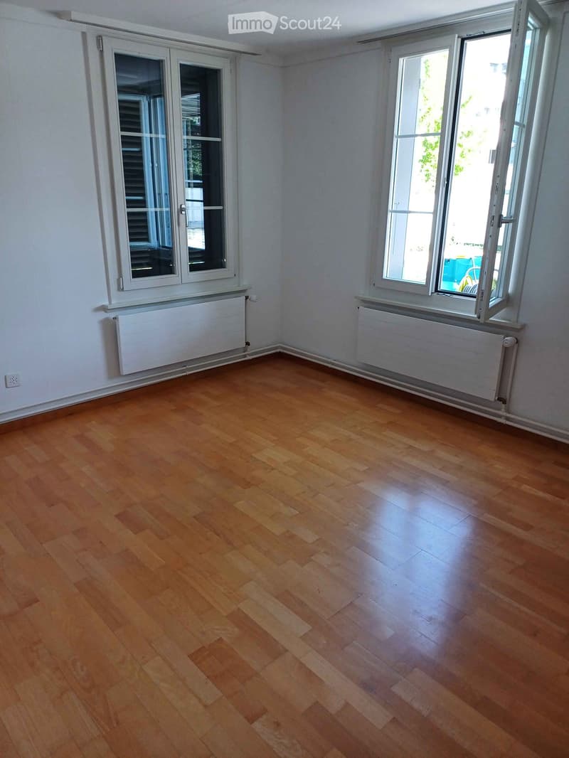 Empty room with wooden flooring, white walls, and large windows with radiators below them.