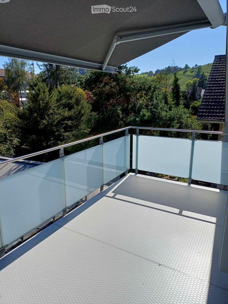 Modern balcony with a glass railing, metal balustrade, and a textured floor, overlooking a lush, green landscape.