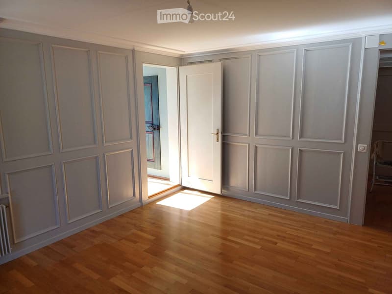 Empty room with wooden flooring and grey walls, featuring a white door and a visible hallway beyond.