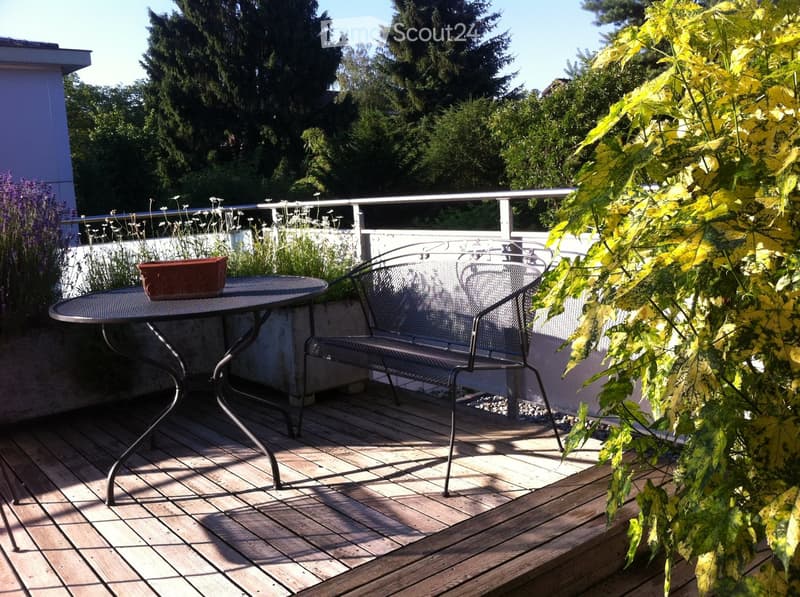 Wooden deck with a round metal table, bench, and flower pot, surrounded by greenery and a metal railing.