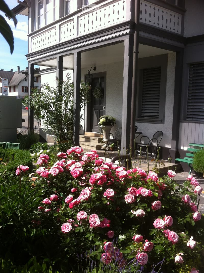 A well-maintained garden with pink roses, lavender, and a small seating area with chairs and a table near the entrance of a house with a decorative balcony.