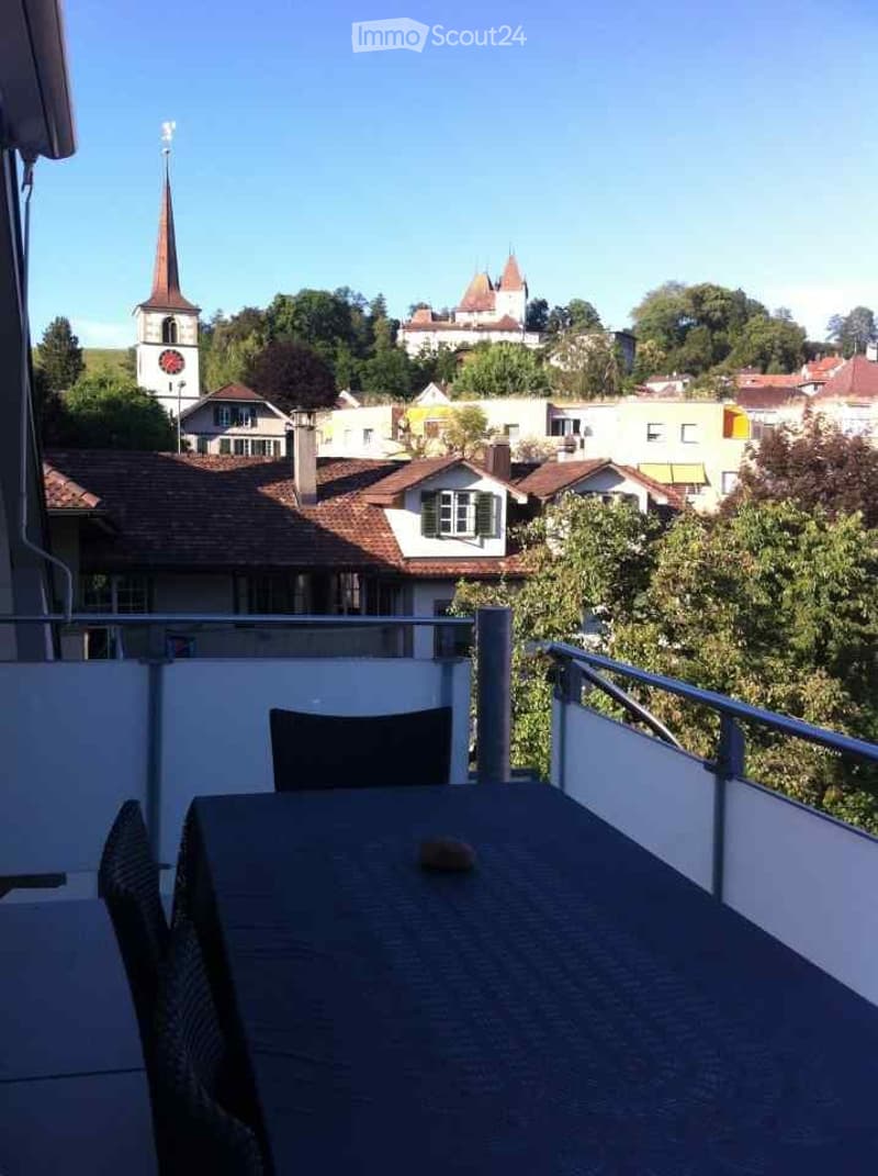Balcony with a table and chair, overlooking a town with a church tower and surrounding houses.