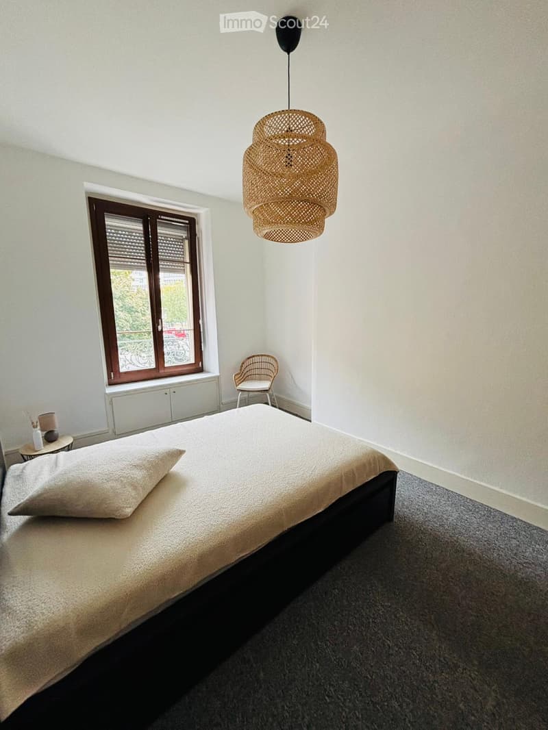A cozy bedroom featuring a double bed with a white bedspread and pillow, a chair next to a window, and a white radiator below it. A small table on the left side has a lamp and a plant. A decorative chandelier is hung from the ceiling.