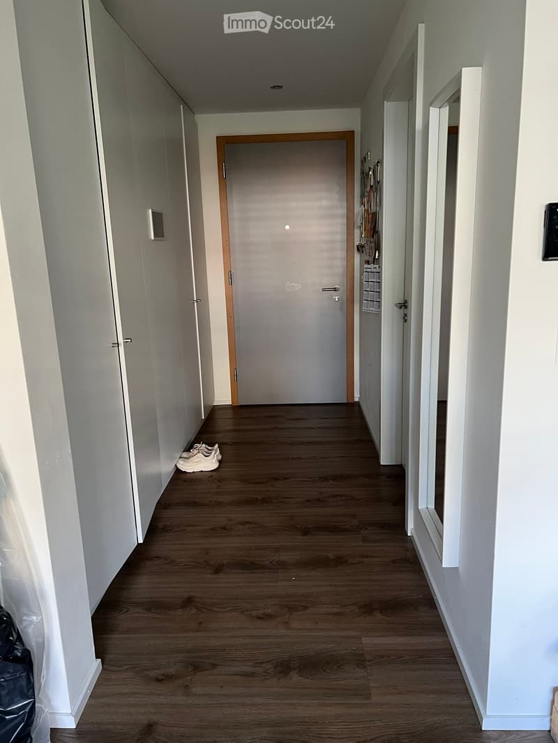 A clean, modern hallway with light wooden flooring, white walls, and a closed door at the end. A pair of white sneakers is placed near the left wall, and a small black bag is visible on the far left side.