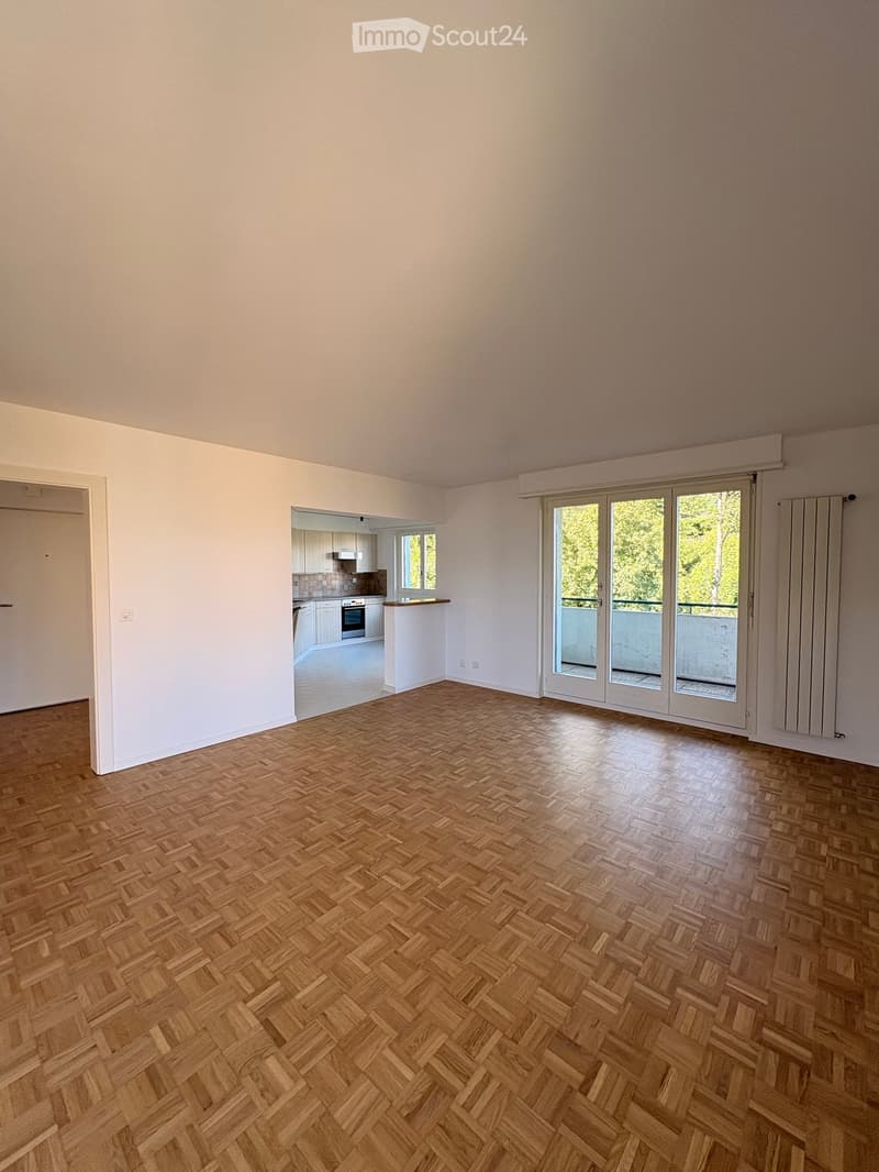 Empty room with wooden herringbone flooring, white walls, and large sliding glass doors leading to a balcony. A small kitchen area is visible in the background.