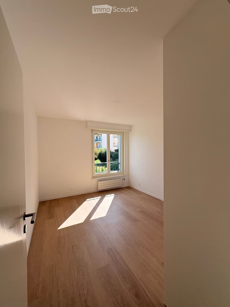 Empty room with wooden flooring, white walls, and a window with a view of a balcony and greenery outside.