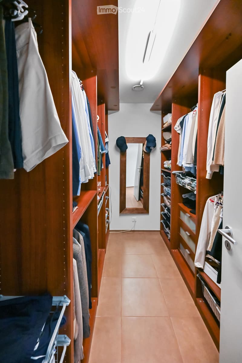 A well-lit hallway with wooden built-in closets on both sides, featuring neatly organized clothing and accessories. The floor is tiled, and a mirror is mounted on the wall at the end of the hallway.