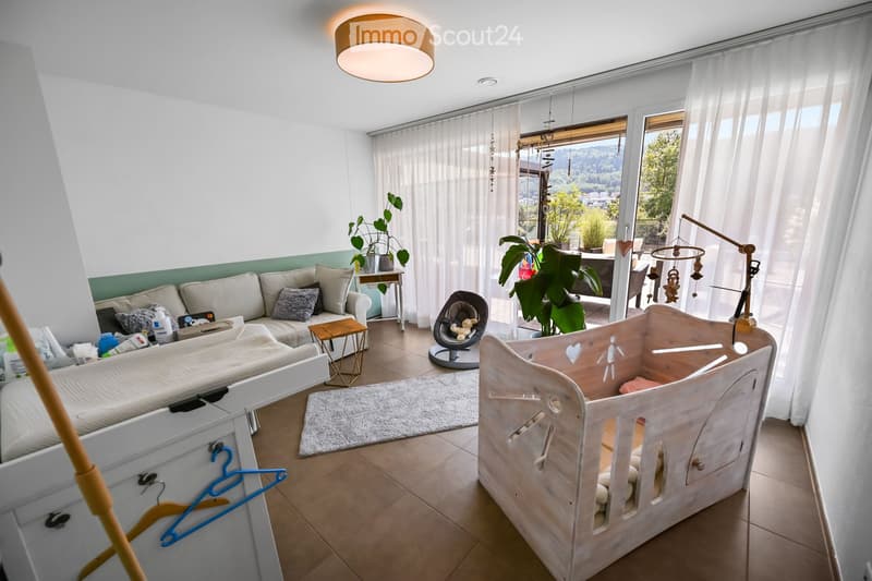 Bright living room with a white sofa, baby crib, baby chair, small table, and plants. Large windows with white curtains lead to a balcony with outdoor furniture.