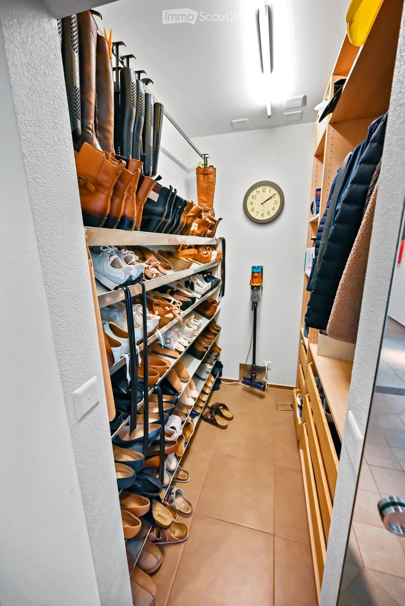 A well-organized hallway with wooden flooring, white walls, and built-in shoe racks filled with various footwear. A clock is mounted on the wall, and a vacuum cleaner is placed near the corner.
