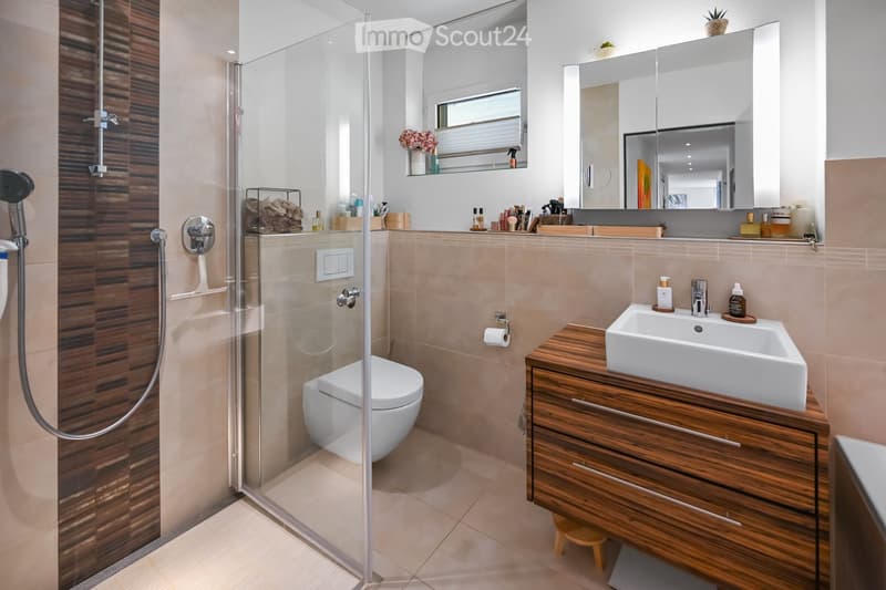 Modern bathroom with a glass-enclosed shower, a toilet, a wooden vanity with a sink, and a large mirror above the sink. The shower features a handheld showerhead and a built-in seat. The vanity has multiple drawers and is topped with a sleek sink. The wal