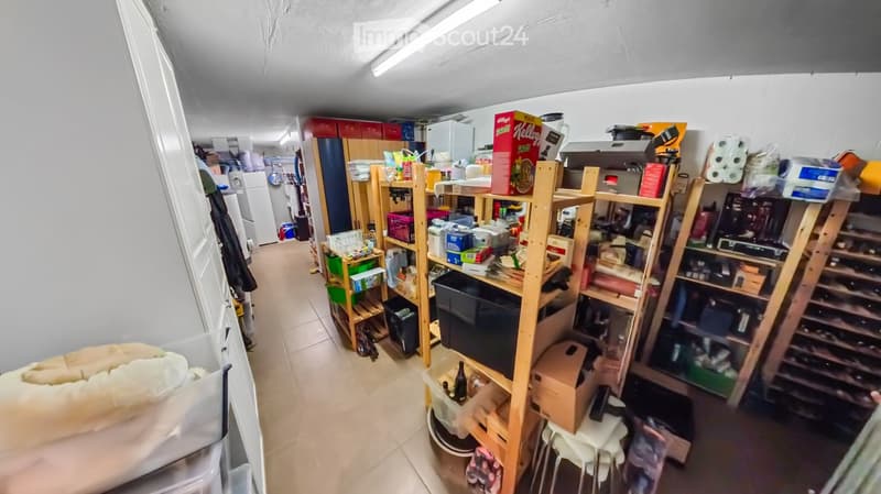 A storage room with wooden shelves filled with various items, including boxes, bottles, and household goods. A refrigerator is visible in the background, along with a wine rack and a small stool.
