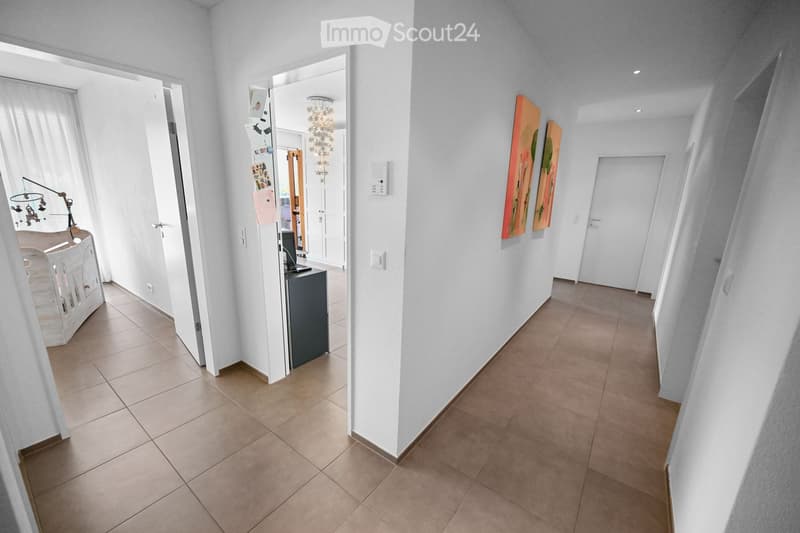 Bright hallway with tiled flooring, white walls, and several doors leading to different rooms. Features include a small table with a lamp, a mirror, and artwork on the walls.