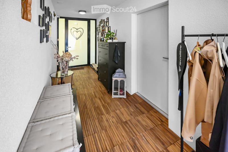 A well-lit hallway with wooden flooring, a white bench, a small round table with a vase, a coat rack with jackets, a black cabinet, and a decorative lantern.