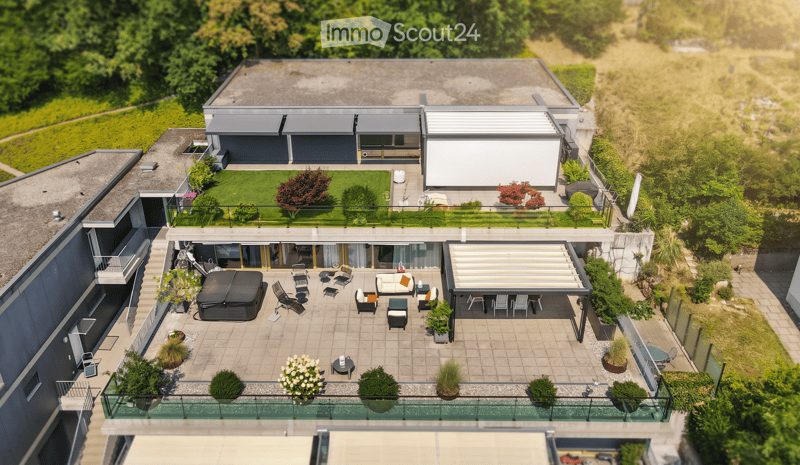 A modern two-story house with a spacious rooftop terrace, featuring lounge chairs, a sofa, a small table, and potted plants. The house has large windows and a sleek design, surrounded by greenery.