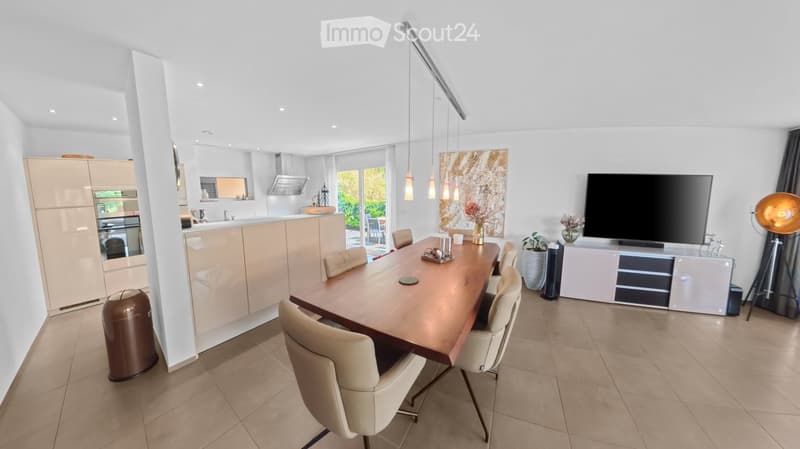 Modern dining area with a wooden dining table, beige chairs, a television, and a kitchen area in the background.