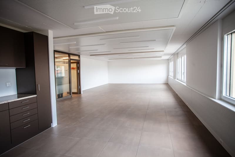 Modern, well-lit corridor with tiled flooring, white walls, and large windows on one side. Features a small kitchenette area with cabinets and a countertop on the left side, and a glass door leading to another room.