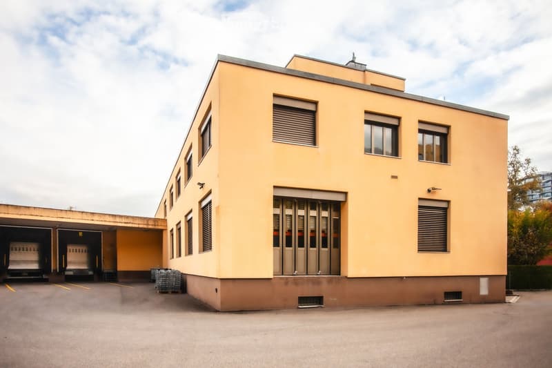 Two-story industrial building with beige facade, large windows, and a loading dock area with pallets.