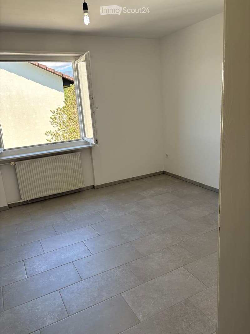 Empty room with tiled floor, white walls, radiator, and large window with view of neighboring building and greenery.