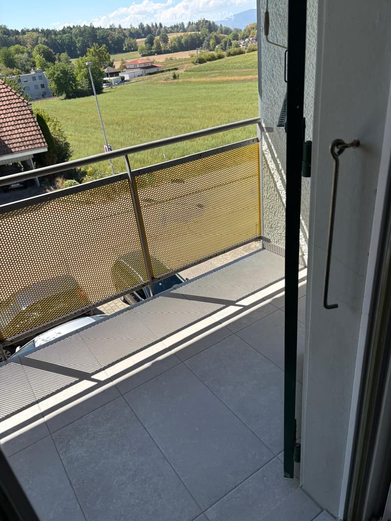 Balcony with metal railing, tiled floor, overlooking a green field and distant mountains.