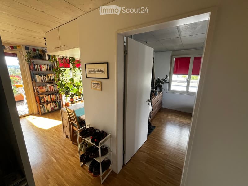 A well-lit hallway with wooden flooring, leading to a room with a bookshelf, dining table, and a shoe rack. The hallway has white walls and a door leading to another room.