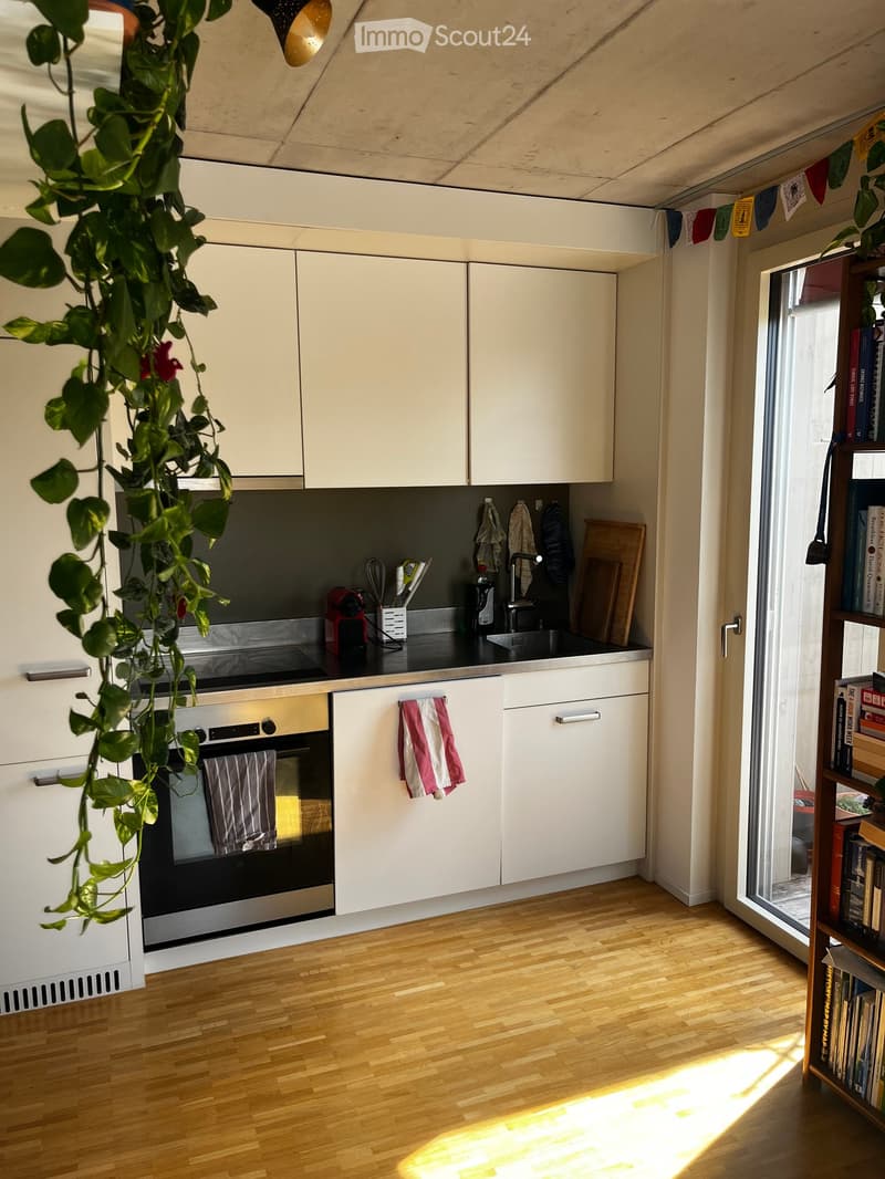 Modern kitchen with white cabinets, wooden flooring, and stainless steel appliances. Features an oven, sink, and a bookshelf with books.
