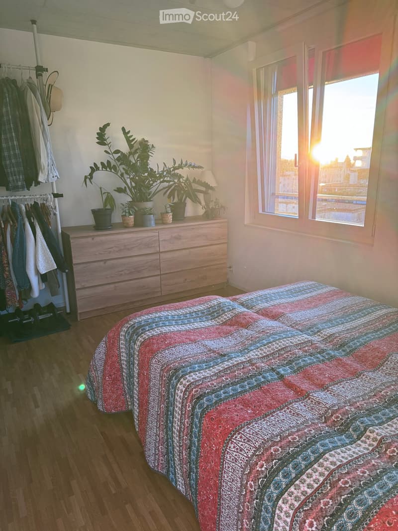 Bright bedroom with wooden flooring, a bed with a colorful quilt, a wooden dresser with potted plants, and a clothing rack with hanging clothes.