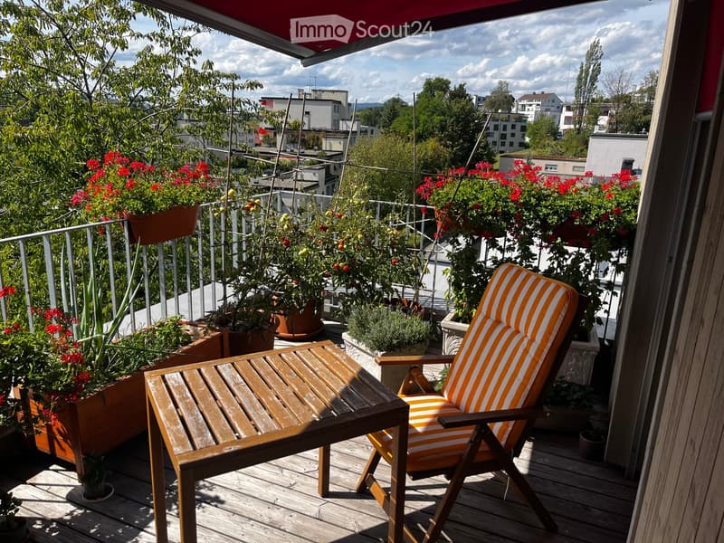 Balcony with wooden decking, potted plants, and outdoor furniture including a wooden table and a striped chair.