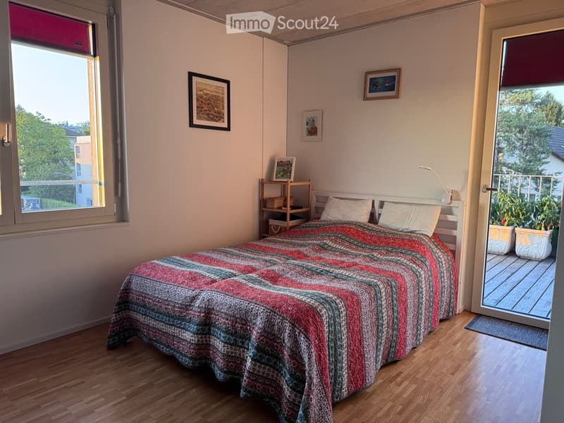 Cozy bedroom with wooden flooring, a neatly made bed with a colorful quilt, a nightstand with a lamp, a small shelf, and framed artwork on the walls. Large windows and a sliding glass door lead to a balcony.