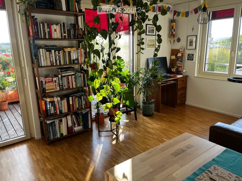 Bright, cozy living space with wooden flooring, large windows, a bookshelf filled with books, a desk with a laptop, and several potted plants. A sofa and coffee table are partially visible.