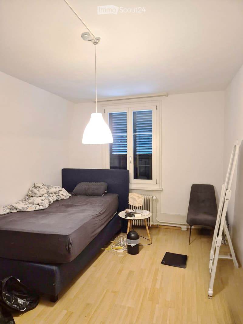 A small bedroom with a single bed, a bedside table, a chair, and a floor lamp. The room has wooden flooring and white walls.
