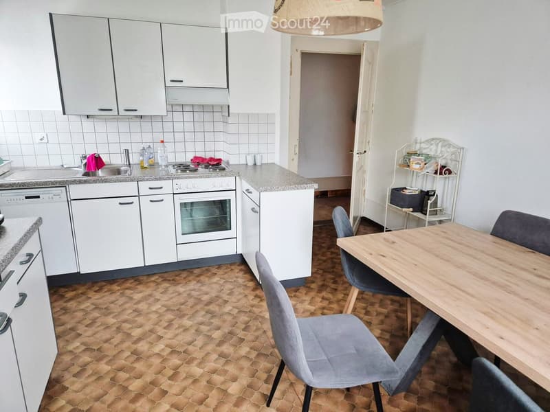 A modern kitchen with white cabinets, a wooden dining table, and grey upholstered chairs. The kitchen features a tiled backsplash, a granite countertop, and a built-in oven and dishwasher.