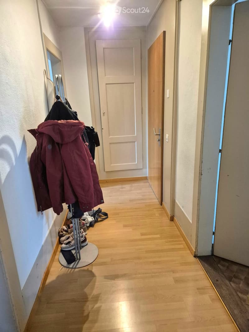 A narrow hallway with wooden flooring, white walls, and a coat rack holding jackets and shoes. A closed door is visible at the end of the hallway.