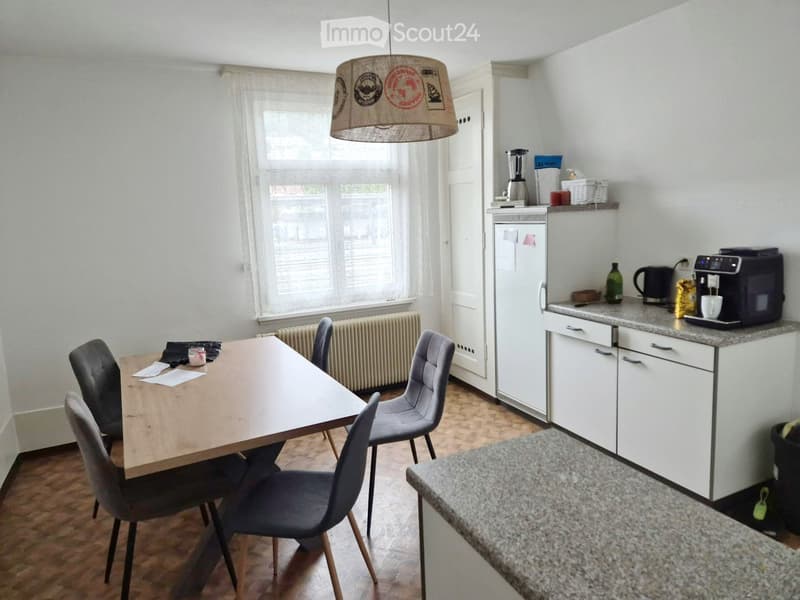 A modern kitchen and dining area with a wooden dining table, four chairs, white cabinets, a refrigerator, coffee machine, and a blender on the counter.