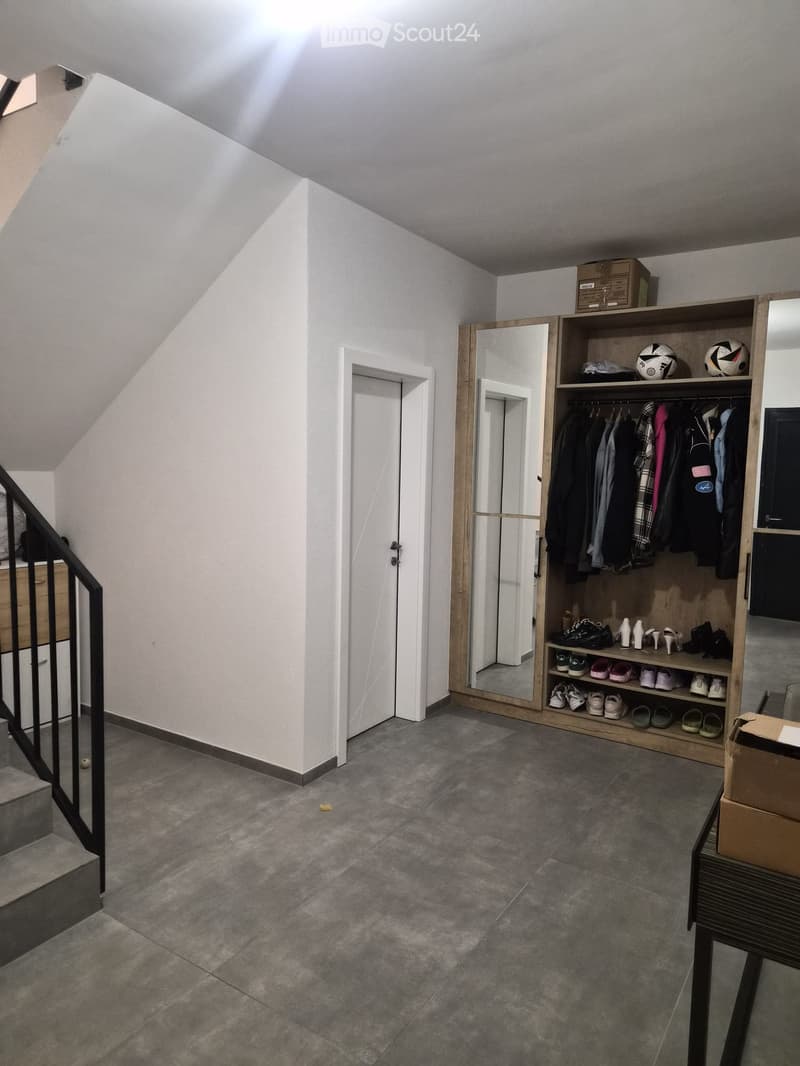 A modern hallway with a staircase, built-in closet, and storage shelves. The space features grey tiled flooring, white walls, and a sleek black metal railing on the staircase.