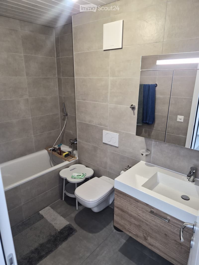 Modern bathroom with a bathtub, toilet, sink, and mirror. Features a wooden cabinet under the sink, a towel on the mirror, and a small shelf next to the bathtub holding toiletries.