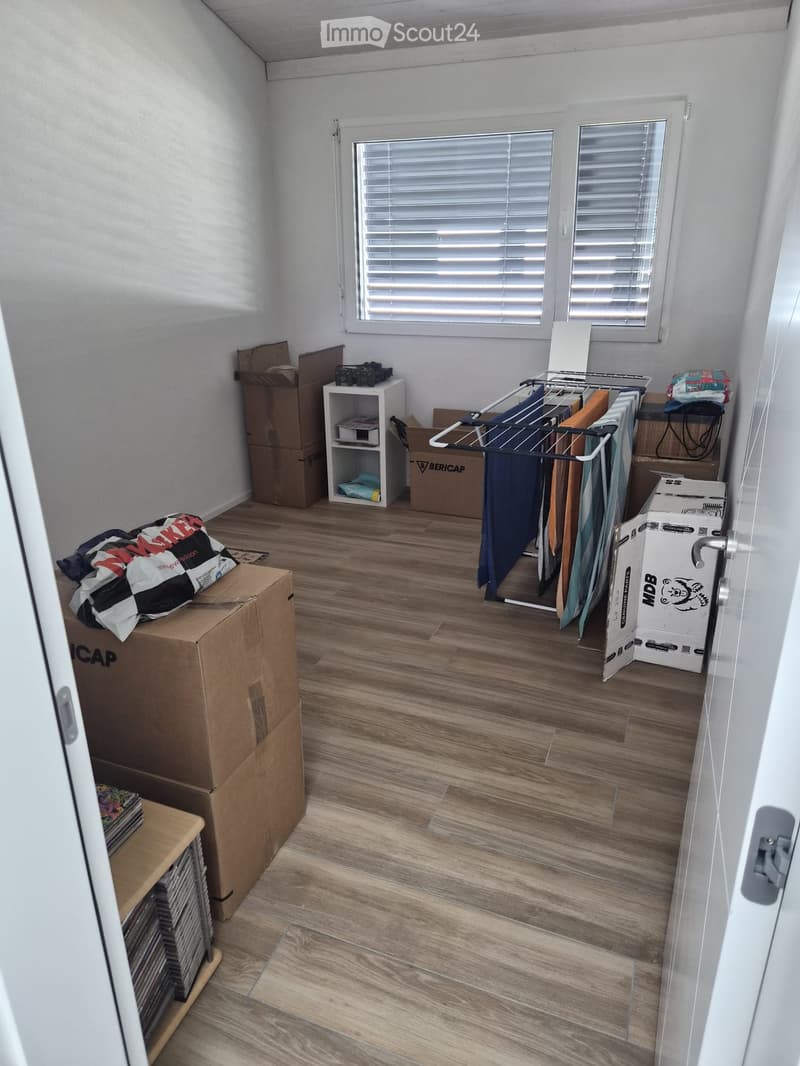 Room with wooden flooring, white walls, and a window with blinds. Contains a clothes drying rack, a small shelf, and several cardboard boxes.