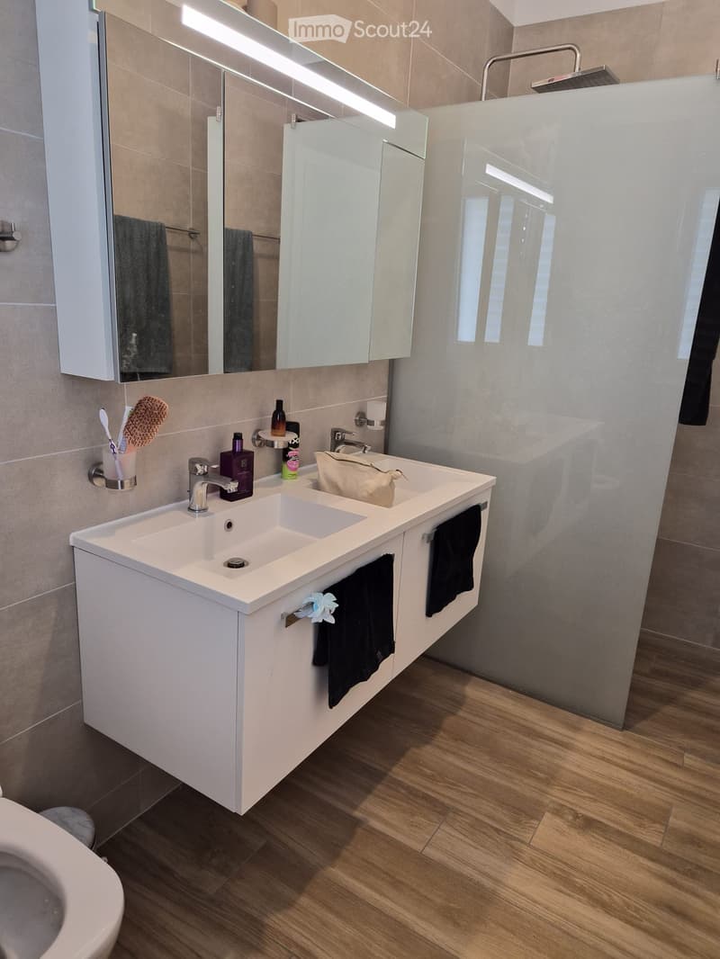 Modern bathroom with double sink, large mirror, shower, and wooden flooring.