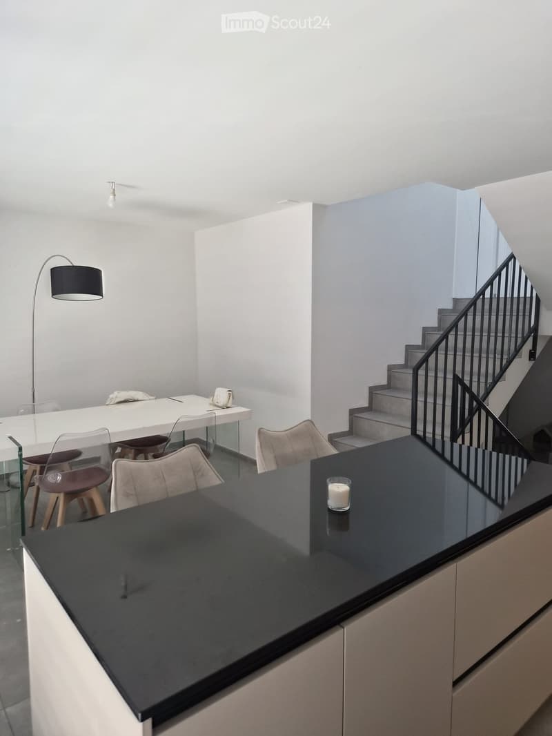 Modern open-plan living and dining area with a sleek black countertop, white dining table, chairs, and a staircase leading upwards.